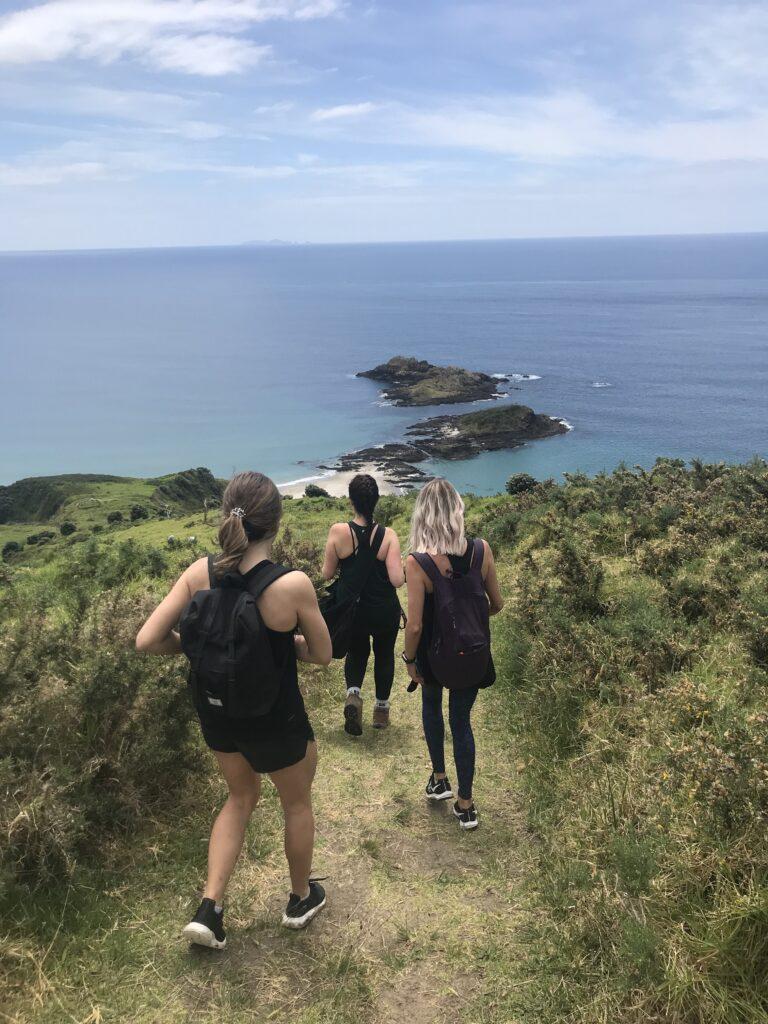 Three women walking Peach Cove track with a view of the bay