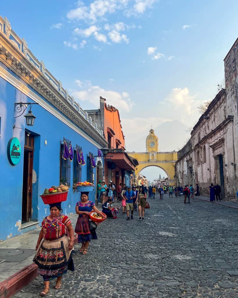 Women carrying fruit baskets on their heads in Antigua, with a landmark and volcano in the distance