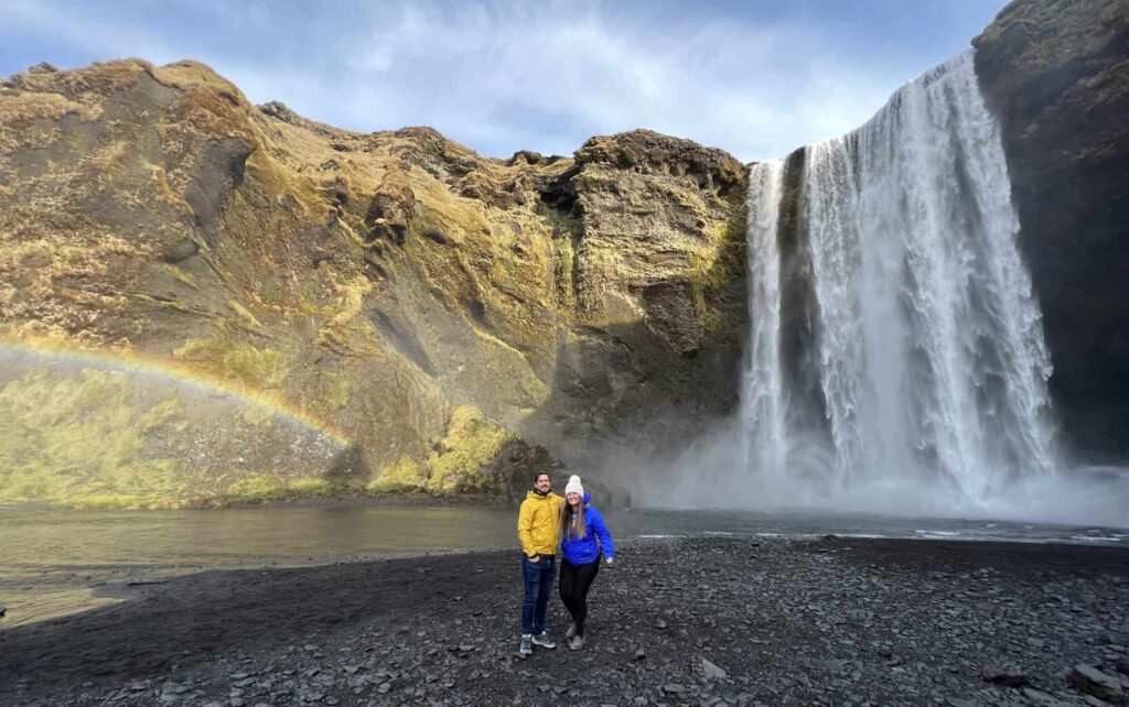 Two people standing between a rainbow and a famous waterfall in Iceland