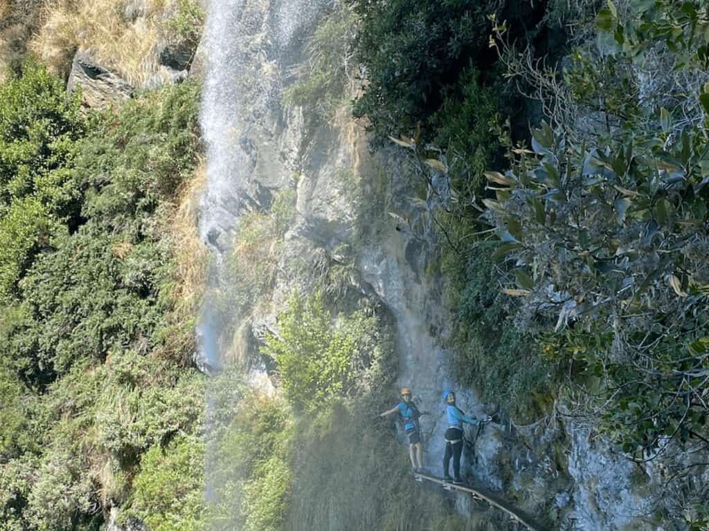 Two women standing on a thin bridge under a waterfall