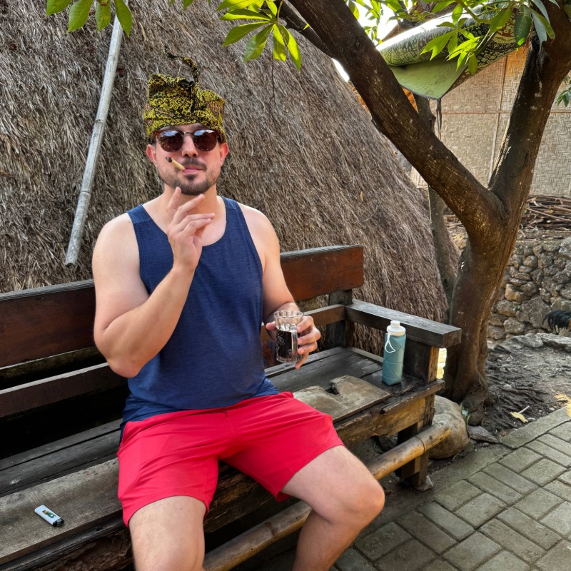 Chesney wearing a traditional hat, smoking tobacco, and drinking Indonesian coffee
