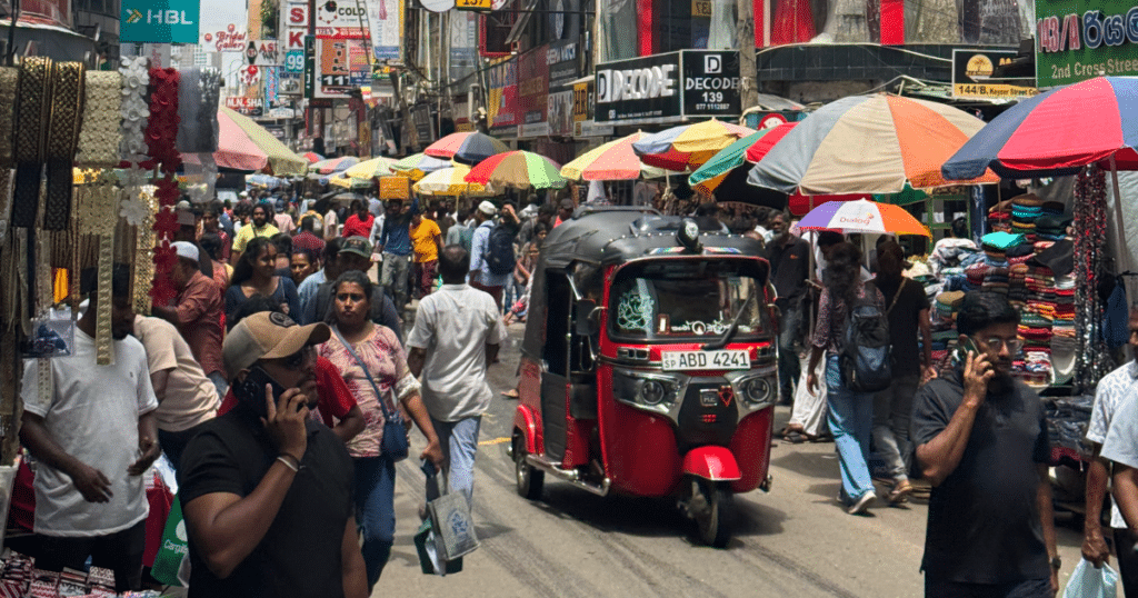 Bustling market street with colorful umbrellas lining stalls and a red auto-rickshaw in the center among shoppers.